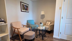Una sala de estar con piso de madera, un sofá, una silla, una mesita con una lámpara y una puerta.
