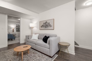 A modern living room with a grey sofa, a round wooden coffee table, a small round side table, and a framed tree artwork on the wall.