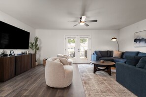 A modern living room with a flat-screen TV, a ceiling fan, a sofa, a coffee table, and a dining area with a wooden cabinet. - Echoes on Eddy - Walk To Stadium & Outdoor Deck! (South Bend)