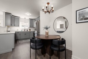 A modern kitchen with grey cabinets, a stainless steel refrigerator, a round dining table with four chairs, and a chandelier.
