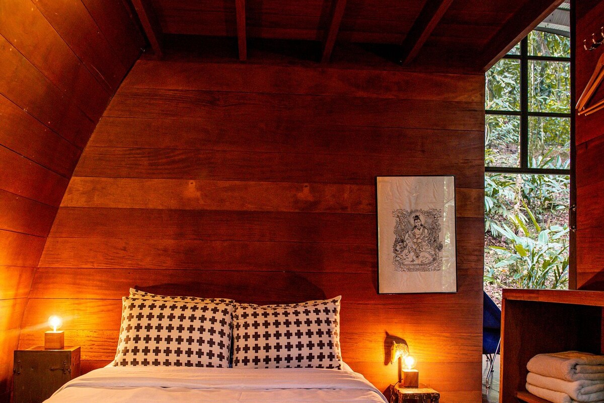 A cozy bedroom with a wooden ceiling, a bed with patterned pillows, a nightstand with a lamp, and a window with a view of greenery.