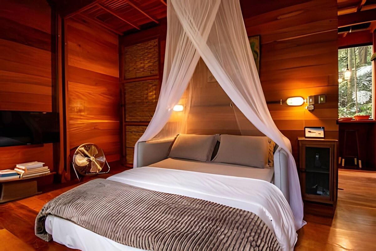 A wooden cabin bedroom with a bed, canopy, bedside lamp, and a nightstand with books.