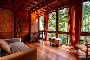 Ein Zimmer mit einem Bett, einem Tisch, Stühlen und Blick auf Grün durch große Fenster.