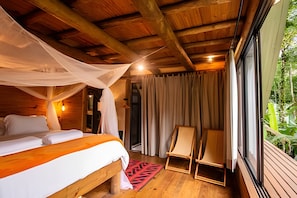 A wooden cabin bedroom with a bed, a chair, and a window overlooking greenery.