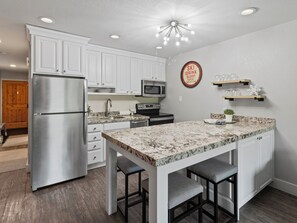 A modern kitchen with white cabinetry, a stainless steel refrigerator, a microwave, and a granite countertop island with bar stools. - Snowcrest 304 | Walk to First Time Lift, Pool & Hot Tub, Mountain Views (Park City)