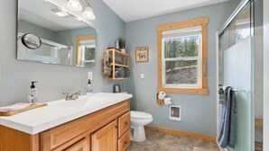 A bathroom with a double sink vanity, a toilet, a large mirror, and a window with a view of the outdoors. - Life's a Bear Haven!!  Couple's Hot Tub under the Sky!  King Bed! (Kalispell)