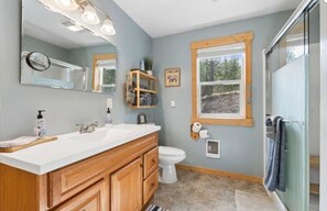 A bathroom with a double sink vanity, a toilet, a large mirror, and a window with a view of the outdoors. - Life's a Bear Haven!!  Couple's Hot Tub under the Sky!  King Bed! (Kalispell)
