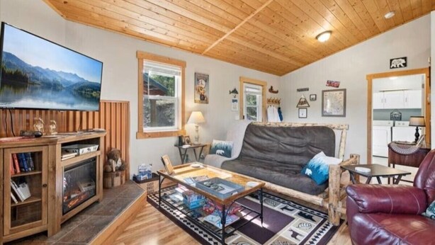 A cozy living room with a wooden ceiling, a flat-screen TV, a fireplace, a sofa, a coffee table, and a bookshelf. - Life's a Bear Haven!!  Couple's Hot Tub under the Sky!  King Bed! (Kalispell)