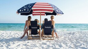 Två strandstolar med etiketten ”Sandestin”, ett parasoll med amerikansk flagga och en strandmiljö.