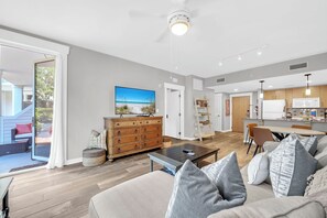 A modern living room with a sofa, coffee table, and a wooden cabinet. There is a television mounted on the wall and a kitchen area in the background. - Resort Amenities-Ground Floor-Walk To Baytowne Wharf-E5109 (Miramar Beach)