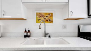 Een keuken met een dubbele spoelbak, witte kasten, een roestvrijstalen kraan, en een lijst met een afbeelding van eten en drinken aan de muur.