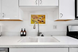 A kitchen with a double sink, white cabinets, a stainless steel faucet, and a framed picture of food and drinks on the wall. - Meiselman Palms - A Ryson Property (Palm Springs)