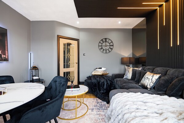 A modern living room with a round table, dark sofas, a large clock, and a door leading to another room.