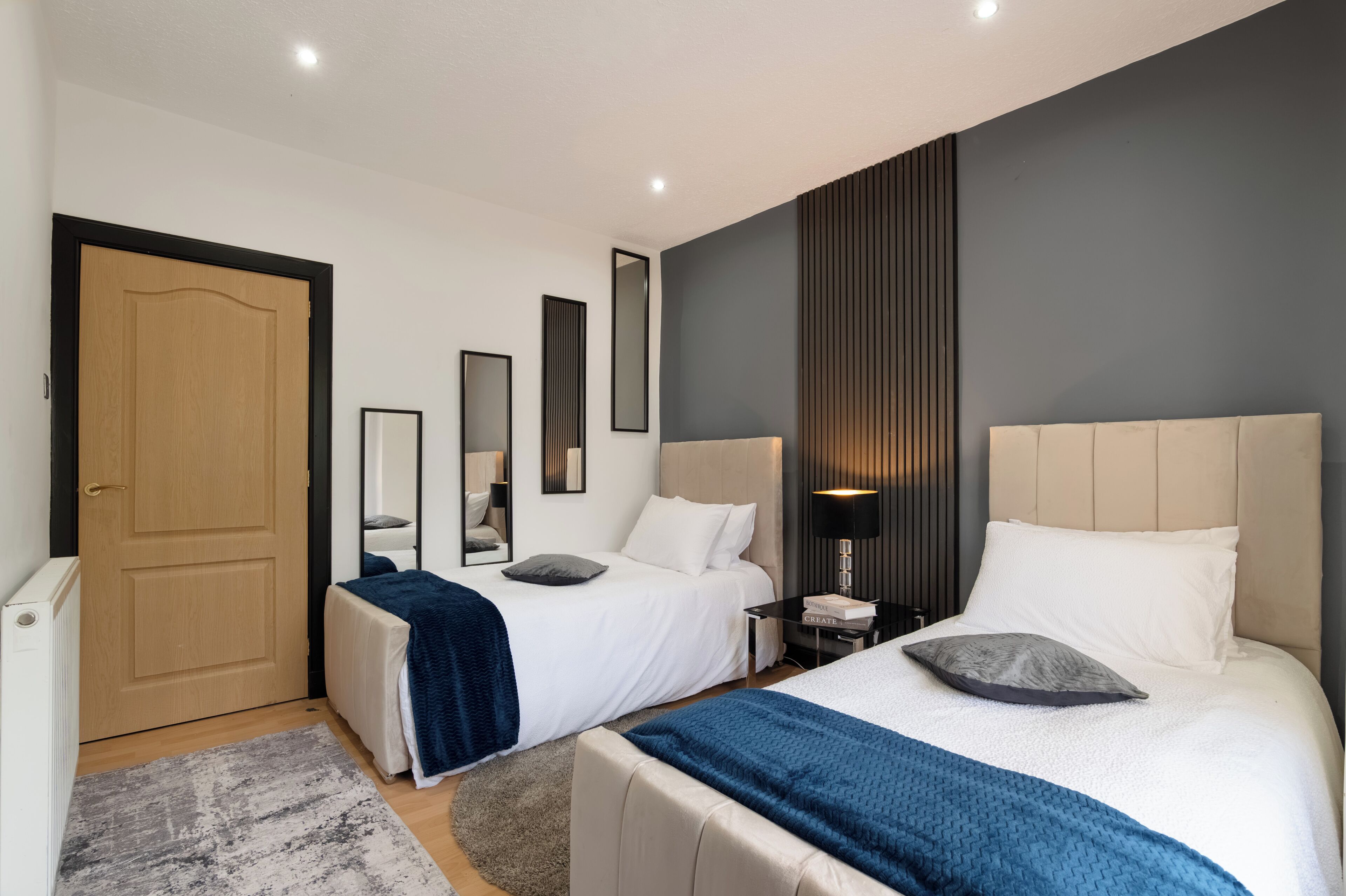 A hotel room with two beds, a bedside table with a lamp, a mirror, and a door.