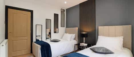 A hotel room with two beds, a bedside table with a lamp, a mirror, and a door.