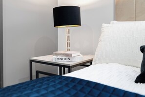 A neatly made bed with a blue quilt, a white pillow, and a black pillow. A small table beside the bed holds a lamp with a black shade and a book titled 'Decoration ideas LONDON'.
