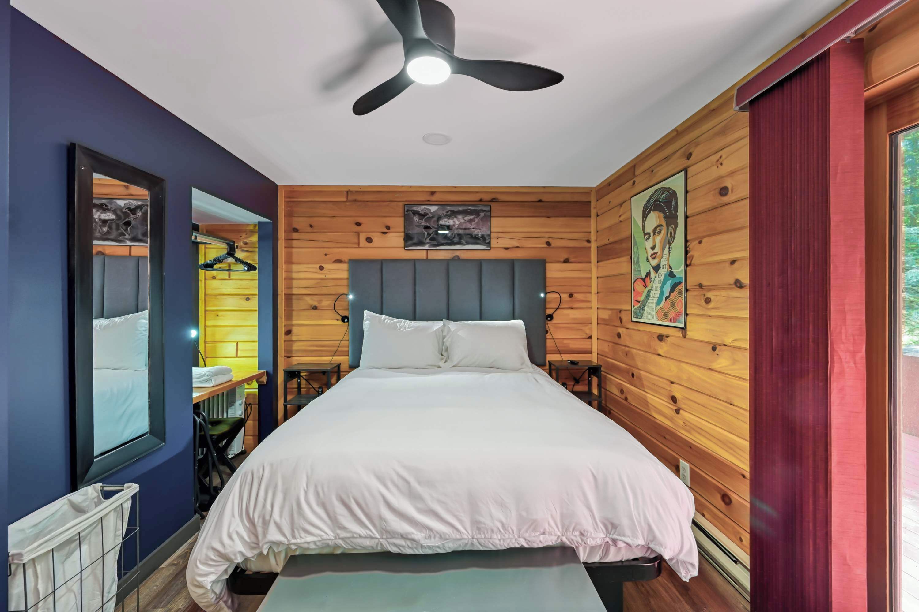A bedroom with a bed, a ceiling fan, wooden walls, a window, and a door.