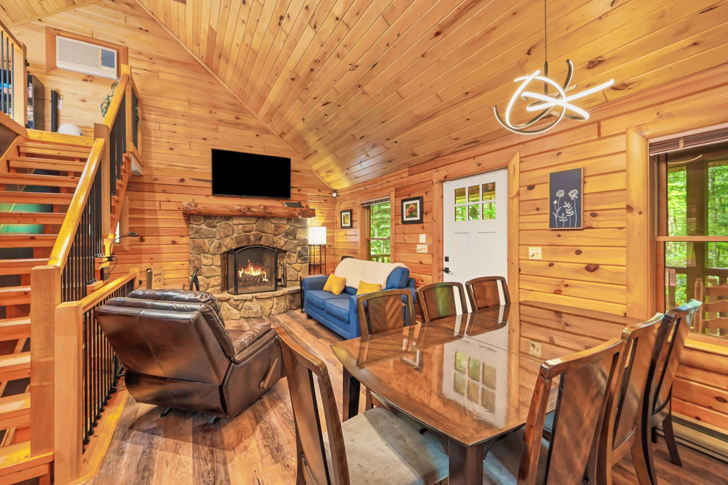 A wooden cabin interior with a stone fireplace, a flat-screen TV, a dining table with chairs, and a staircase.