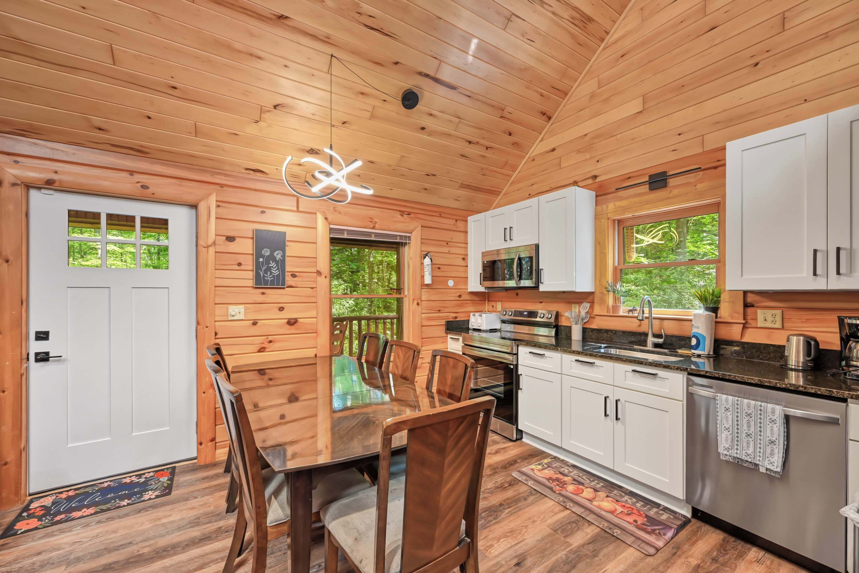 A wooden interior with a dining table, chairs, a kitchen with appliances, and a view of greenery outside.