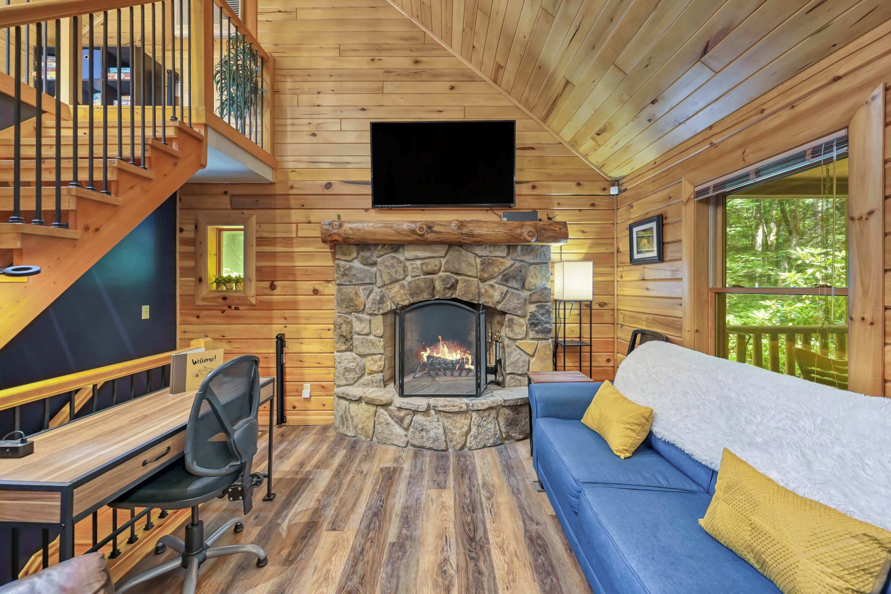 A cabin interior with a stone fireplace, a blue sofa, a wooden desk, and a staircase.