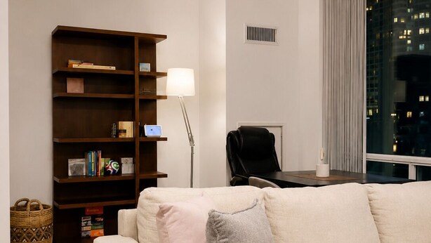 Ett modernt vardagsrum med en beige soffa, en bokhylla i trä, en golvlampa och utsikt över en stadsbild genom ett fönster.
