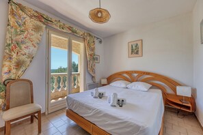 Ein Schlafzimmer mit Bett, einem Sessel, einem Nachttisch mit Lampe, einem Fenster mit Vorhängen und einem Wandbild.