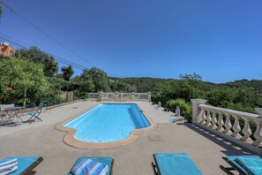 Ein rechteckiger Pool mit gewölbter Kante, umgeben von einer Betonplattform. Liegestühle mit Handtüchern, ein Zaun und ein Blick auf eine hügelige Landschaft sind zu sehen.