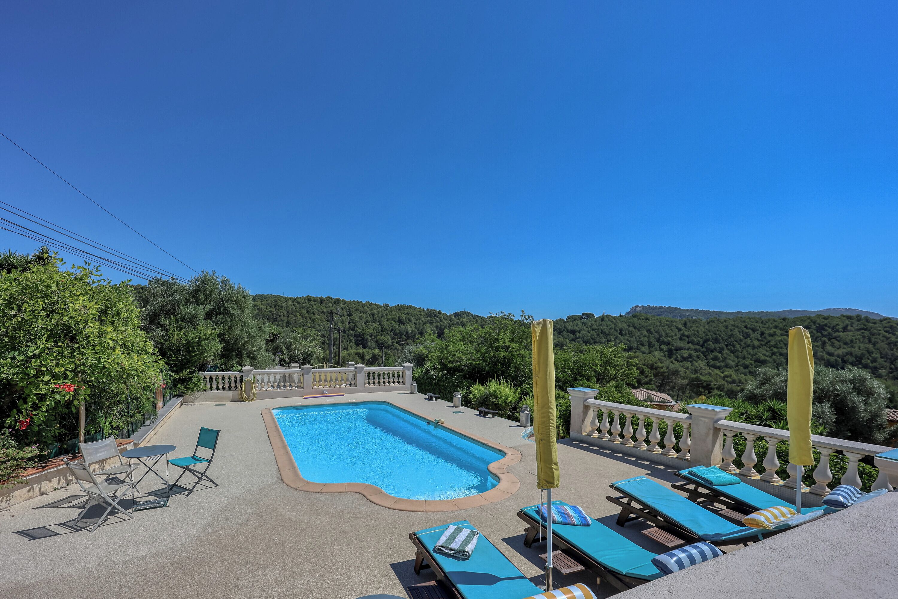 Une piscine entourée d’une clôture et de verdure, avec des chaises longues et une table à proximité.