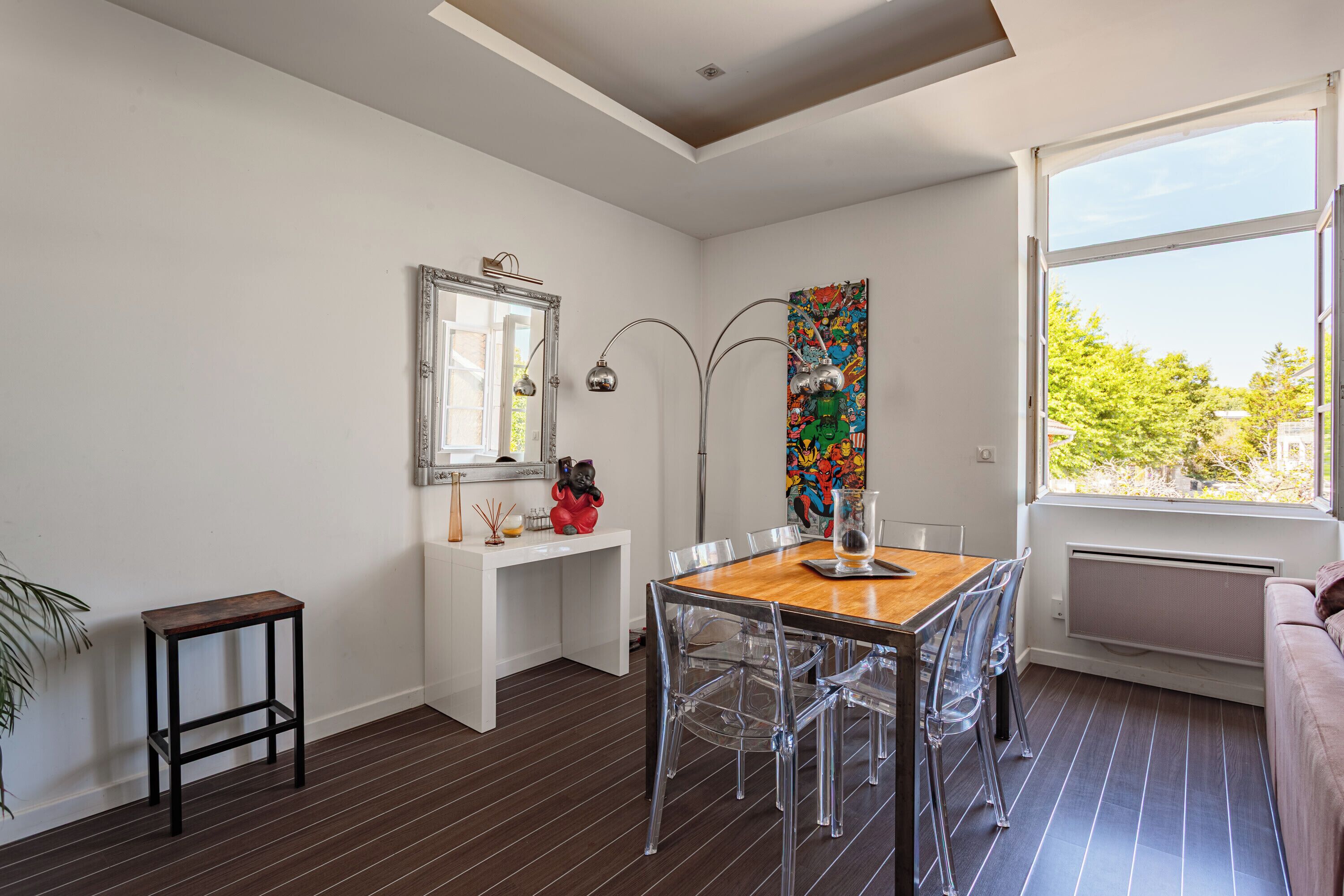A modern dining area with a wooden table and clear chairs, a white console table with decorative items, a large mirror, and a colorful artwork on the wall.