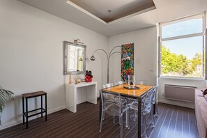 Ein moderner Essbereich mit einem Holztisch und transparenten Stühlen, einem weißen Konsolentisch mit Dekorationsobjekten, einem großen Spiegel und einem farbenfrohen Kunstwerk an der Wand.