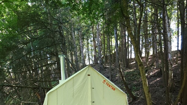 Ein Zelt mit einer Rampe, zwei Stühlen, einer Mülltonne und einem Wagen in einem Wald.