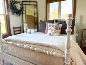 A bed with a white headboard, a floral pillow, and a plain bedspread in a room with a window and a mirror.