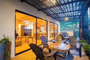 Un espace repas extérieur moderne, doté de chaises en bois et d’une table ronde, et relié à un espace intérieur par une porte vitrée.