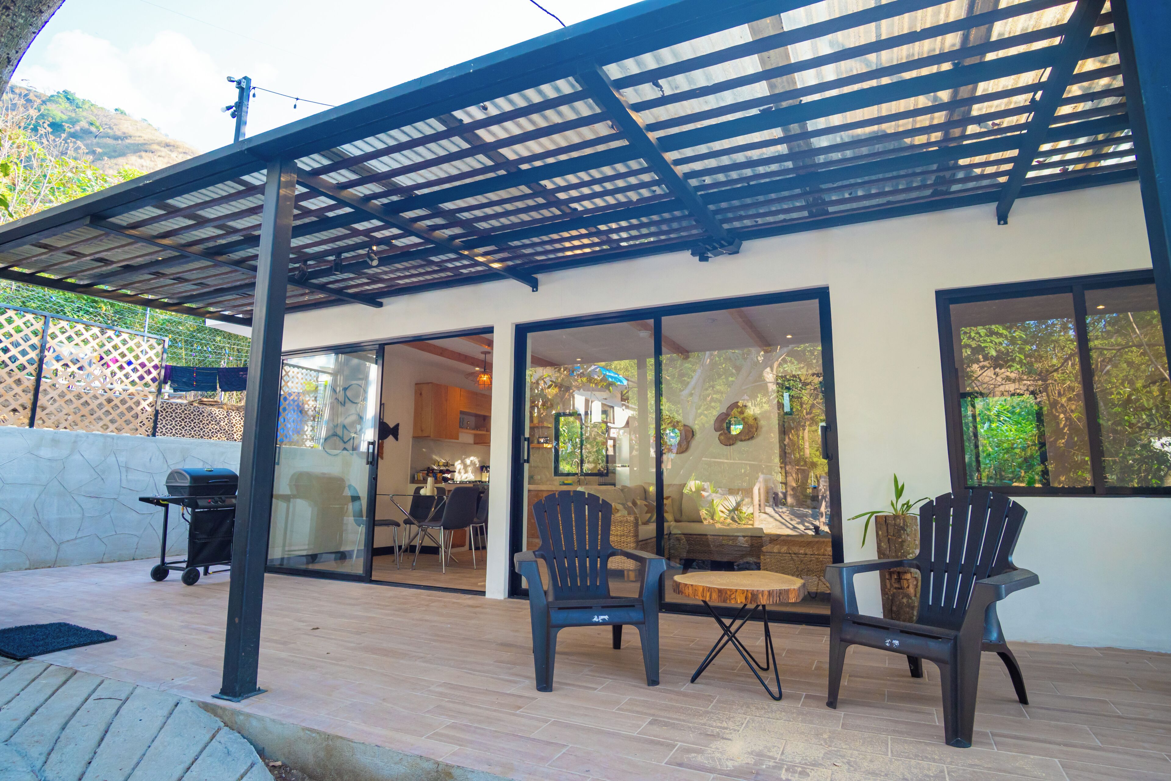 An outdoor patio with a pergola, two Adirondack chairs, a round wooden table, and a grill.
