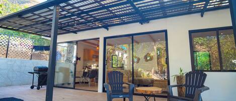 An outdoor patio with a pergola, two Adirondack chairs, a round wooden table, and a grill.
