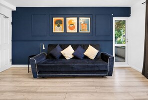 Une pièce aux murs bleu foncé, avec un canapé noir, trois œuvres d’art encadrées et une porte donnant sur un balcon.