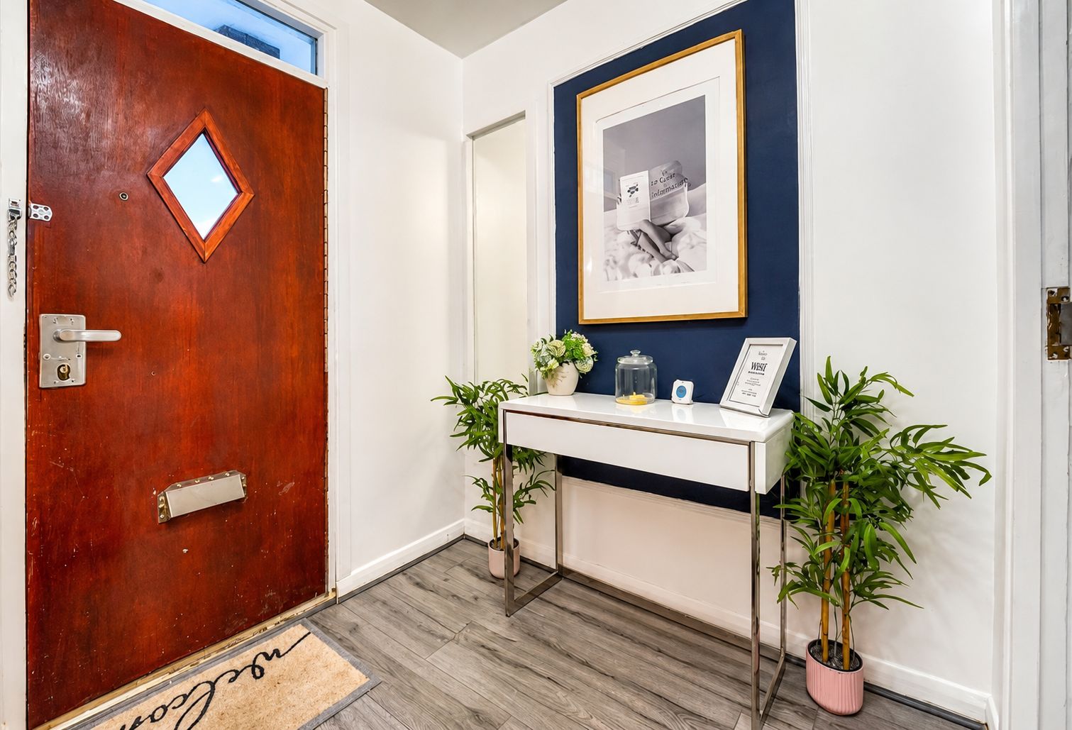 Uma porta de madeira com uma pequena janela, um console branco com um quadro emoldurado e duas plantas em vasos.
