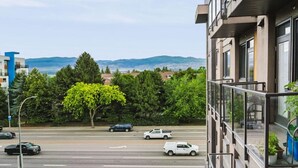 A view from a balcony overlooking a street with cars, trees, and distant mountains. - The Knox | Central Pet-Friendly Kelowna 2BR Condo (Kelowna)