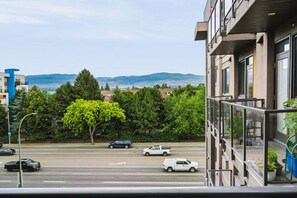 A view from a balcony overlooking a street with cars, trees, and distant mountains. - The Knox | Central Pet-Friendly Kelowna 2BR Condo (Kelowna)
