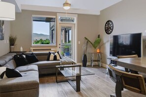 A modern living room with a sofa, coffee table, and a TV. There is a dining area with a table and chairs. A large window provides a view of the outdoors. - The Knox | Central Pet-Friendly Kelowna 2BR Condo (Kelowna)