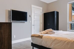 A bedroom with a bed, a television mounted on the wall, a wardrobe, and a window. - The Knox | Central Pet-Friendly Kelowna 2BR Condo (Kelowna)