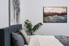 A bedroom with a bed, a nightstand, a potted plant, and two artworks on the wall. - The Knox | Central Pet-Friendly Kelowna 2BR Condo (Kelowna)