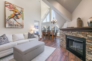A living room with a fireplace, a sofa, an ottoman, and a dining area with a chandelier. - True Ski In/ Out 3 BD Lodge at Mtn Village, Pool, Hot Tub, Fitness (Park City)