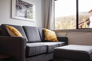 Ein graues Sofa mit zwei gelben Kissen, ein gerahmtes Bild an der Wand und ein Fenster mit Blick auf eine bewaldete Landschaft.