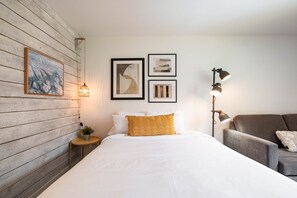 Ein Schlafzimmer mit einer Holzwand, einem Bett mit weißen und senffarbenen Kissen, einem grauen Sofa und gerahmten Kunstwerken.