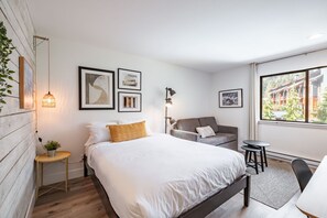 A modern bedroom with a large bed, a sofa, a small table, and a view of a building through a window.
