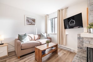 A modern living room with a sofa, a coffee table, a TV mounted on the wall, and a fireplace. - Creekside | Walk to Gondola | Quiet Studio (Whistler)