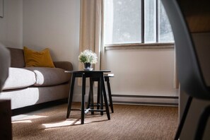 A living room with a sofa, a small table with a plant, and a window with blinds. - Creekside | Walk to Gondola | Quiet Studio (Whistler)