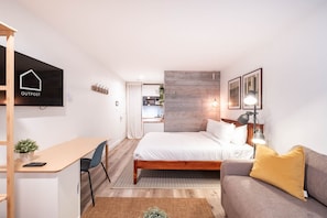 A compact hotel room with a bed, a desk, a sofa, a TV, and a kitchenette.