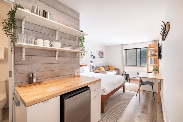 A compact hotel room with a kitchenette, a bed, a desk, and a window.